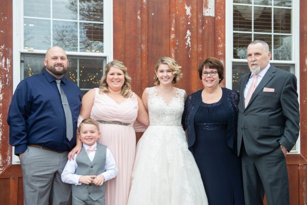 Christine-Jon-Wedding-Blog-44 - TOMMY GATZ ENTERTAINMENT Group portrait with six people in formal attire, including a young boy and a woman in a wedding dress, standing in front of a rustic building with large windows.