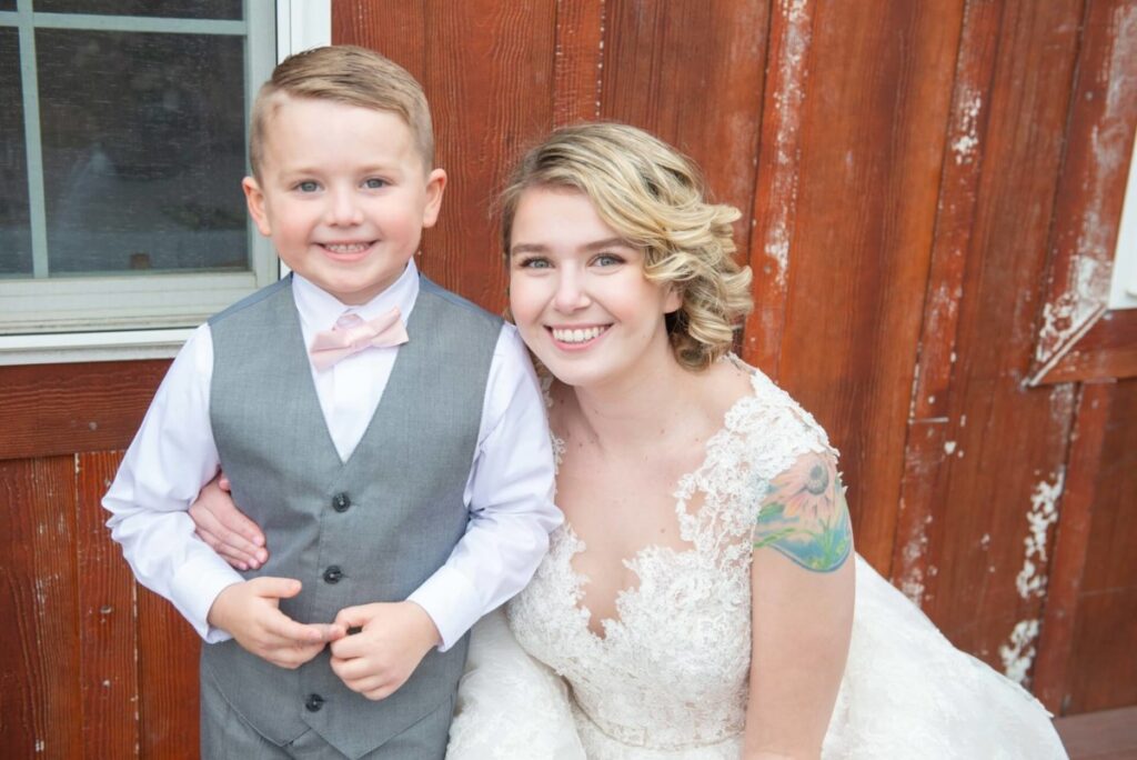 Christine-Jon-Wedding-Blog-45 - TOMMY GATZ ENTERTAINMENT A young woman in a white lace dress and a child in a gray vest with a bow tie smile in front of a wooden wall.