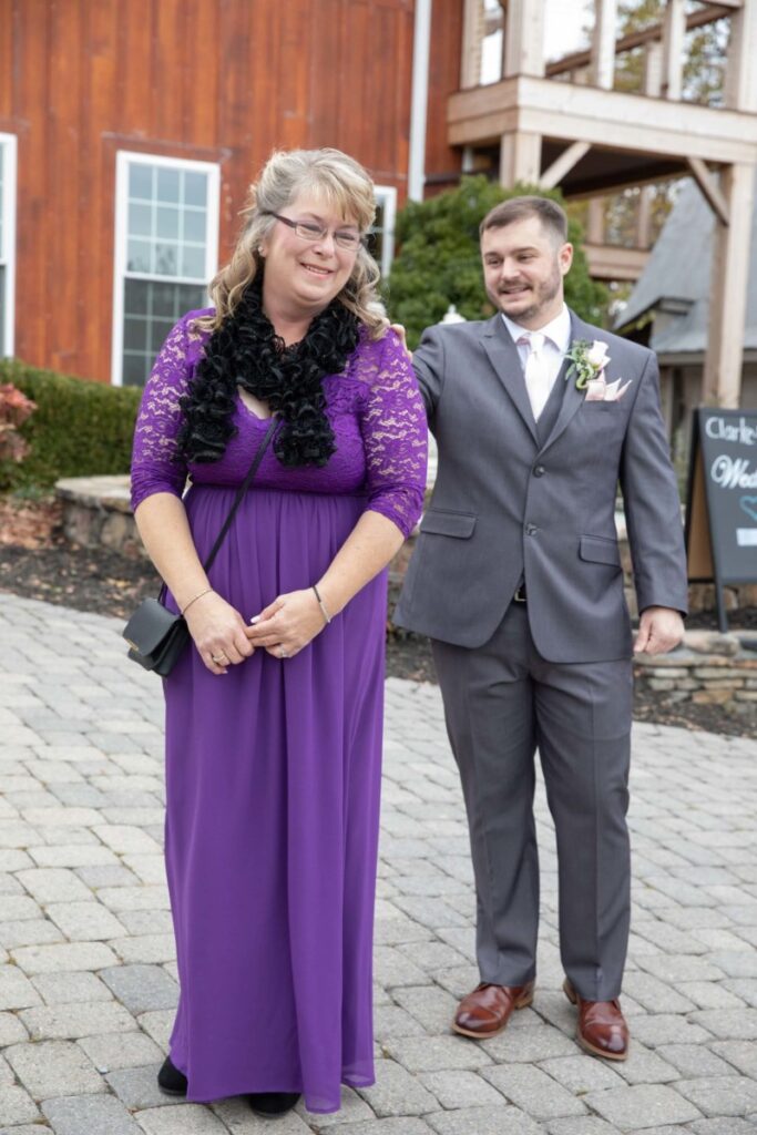 Christine-Jon-Wedding-Blog-46 - TOMMY GATZ ENTERTAINMENT A woman in a purple lace dress and a man in a gray suit stand outdoors on a paved walkway near a wooden building.