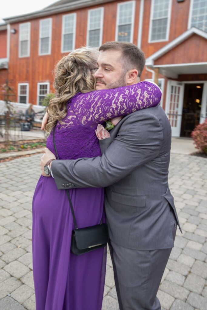 Christine-Jon-Wedding-Blog-47 - TOMMY GATZ ENTERTAINMENT A man in a gray suit hugs a woman in a purple dress outside a red building on a stone path.