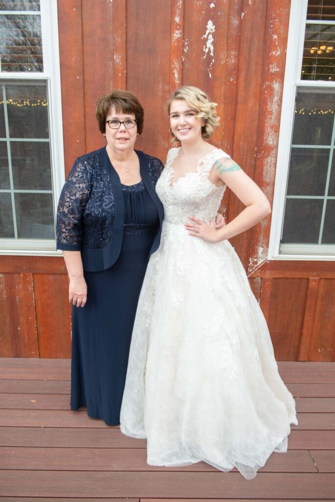 Christine-Jon-Wedding-Blog-48 - TOMMY GATZ ENTERTAINMENT Two women stand in front of a wooden wall, one in a formal blue dress and the other in a white lace gown, smiling and posing for a photo.