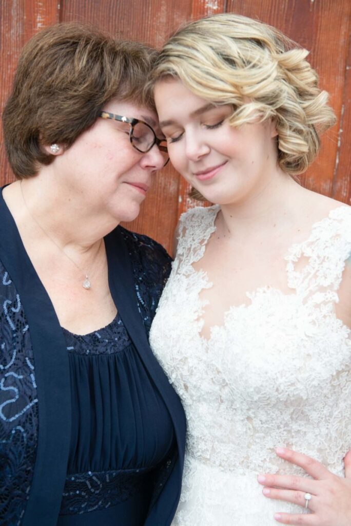 Christine-Jon-Wedding-Blog-49 - TOMMY GATZ ENTERTAINMENT Two women standing closely, with eyes closed, in front of a wooden background. One is in a lace dress, the other in a blue outfit. They appear to share a tender moment.