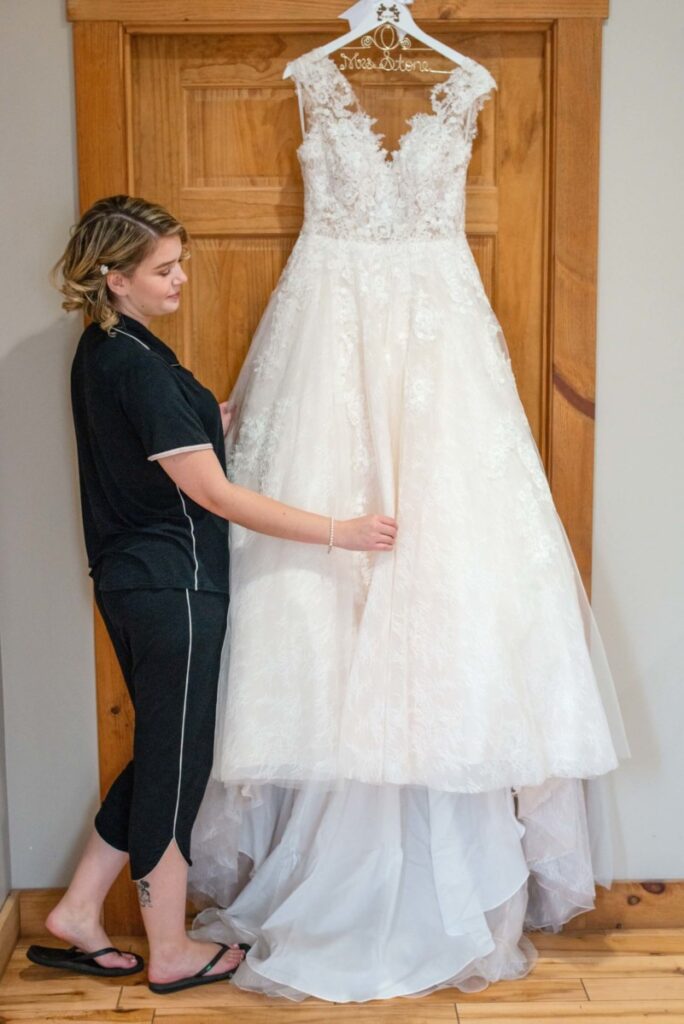 Christine-Jon-Wedding-Blog-5 - TOMMY GATZ ENTERTAINMENT Person in casual attire holding a white lace wedding dress hanging on a wooden door.