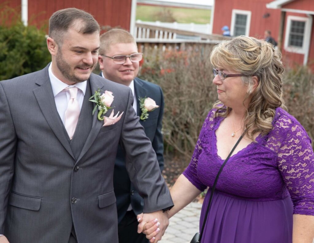 Christine-Jon-Wedding-Blog-50 - TOMMY GATZ ENTERTAINMENT A man in a suit holding hands with a woman in a purple dress outdoors. Another man in a suit is in the background. They stand on a paved walkway with red buildings and shrubs nearby.