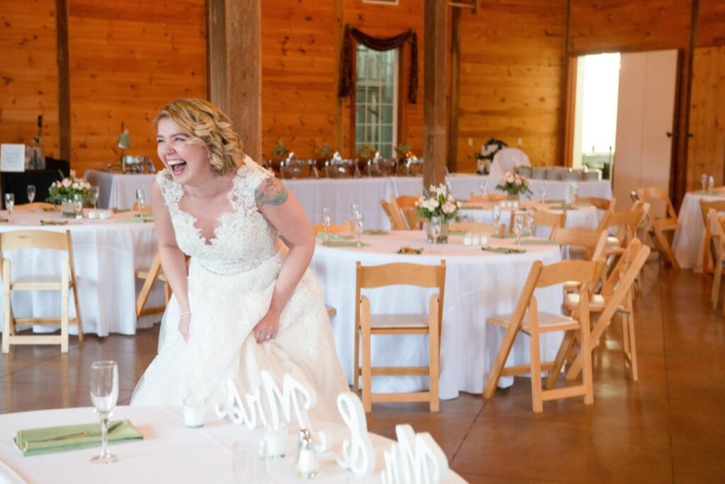 Christine-Jon-Wedding-Blog-51 - TOMMY GATZ ENTERTAINMENT Bride in a white gown laughing joyfully in a rustic wedding venue with wooden chairs and white tablecloths.