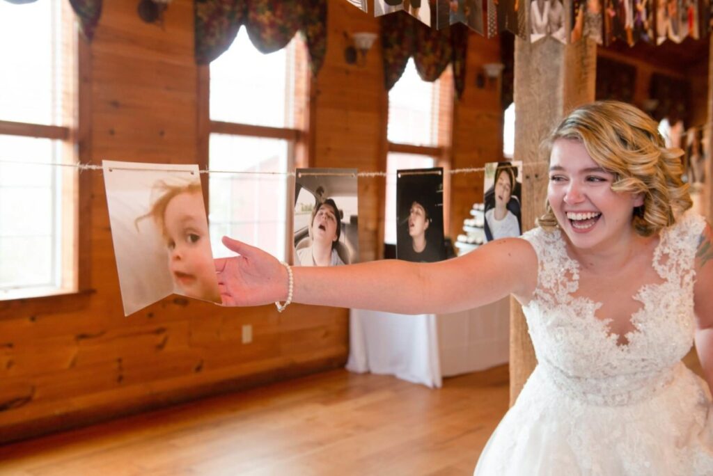Christine-Jon-Wedding-Blog-52 - TOMMY GATZ ENTERTAINMENT A bride in a lace dress smiles and holds up a string of photographs indoors, featuring various expressions of a child's face.