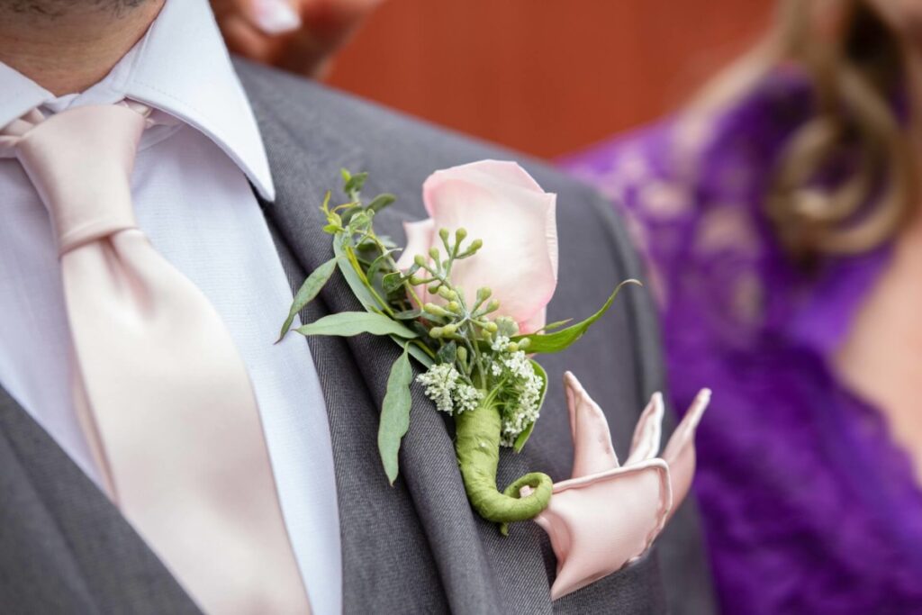 Christine-Jon-Wedding-Blog-53 - TOMMY GATZ ENTERTAINMENT Close-up of a man in a gray suit, wearing a pink tie and pocket square, with a boutonniere featuring a pink rose and greenery. A person in a purple dress is partially visible in the background.