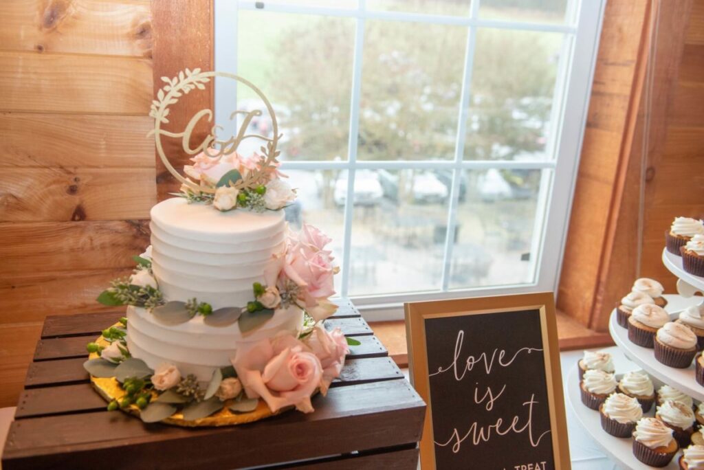 Christine-Jon-Wedding-Blog-55 - TOMMY GATZ ENTERTAINMENT A two-tier white cake with pink roses and a circular topper on a wooden table. A sign next to it reads "Love is sweet." Cupcakes are displayed on the right.