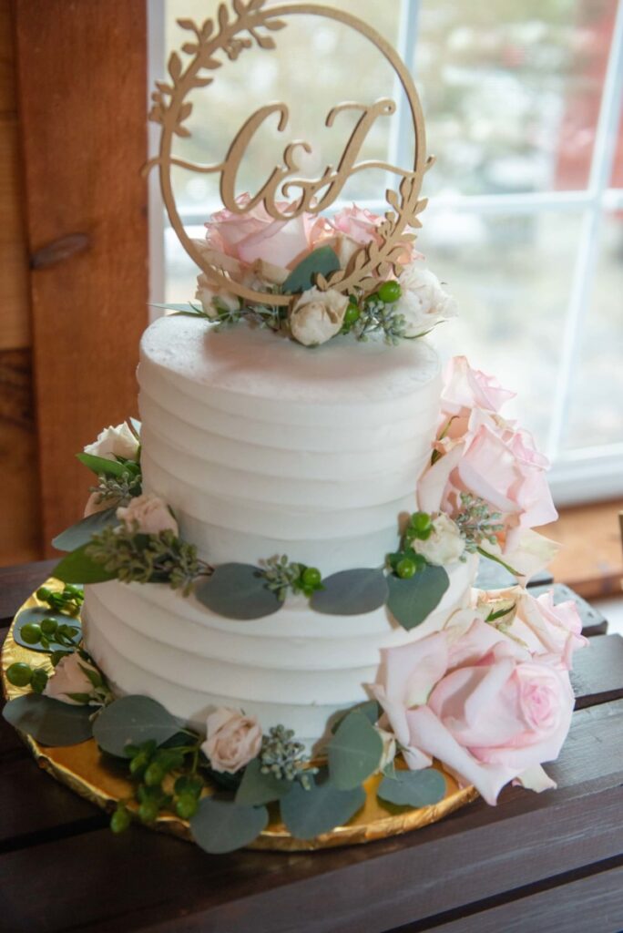 Christine-Jon-Wedding-Blog-56 - TOMMY GATZ ENTERTAINMENT Two-tier white frosted cake adorned with pink roses, greenery, and a gold monogram topper on a wooden table.