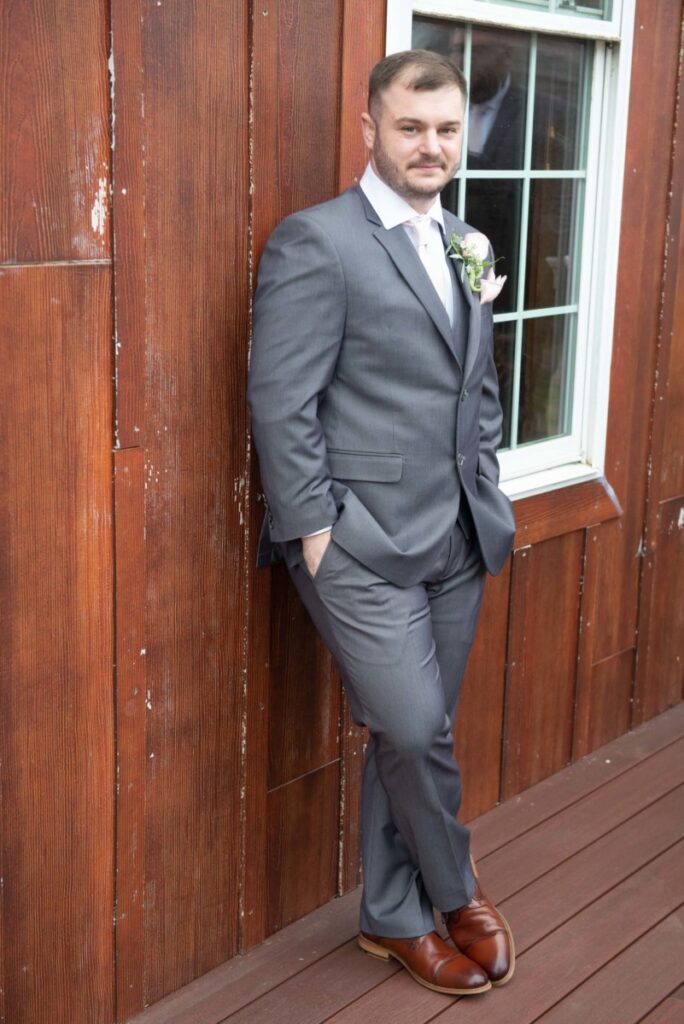 Christine-Jon-Wedding-Blog-58 - TOMMY GATZ ENTERTAINMENT Man in gray suit and brown shoes leans against a wooden wall next to a window, with hands in pockets.