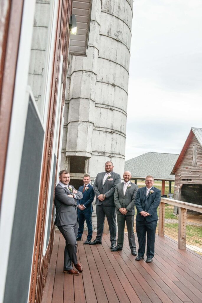 Christine-Jon-Wedding-Blog-59 - TOMMY GATZ ENTERTAINMENT Five men in suits pose on a wooden deck beside a tall, cylindrical structure and rustic buildings.