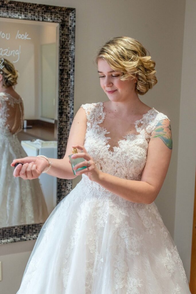 Christine-Jon-Wedding-Blog-6 - TOMMY GATZ ENTERTAINMENT A woman in a lacy white wedding dress is applying perfume on her wrist. She stands in front of a decorative mirror with her reflection visible.
