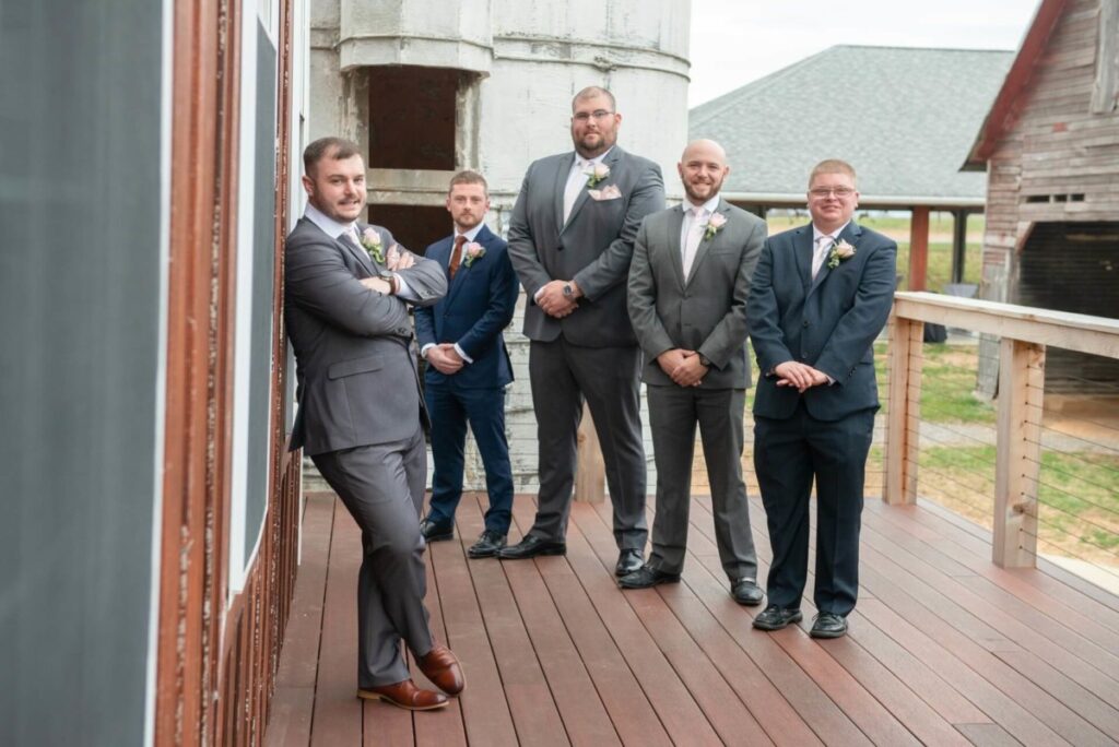Christine-Jon-Wedding-Blog-60 - TOMMY GATZ ENTERTAINMENT Five men in suits stand on a wooden deck; one leans against a wall while the others stand in a row, posing for a photo.