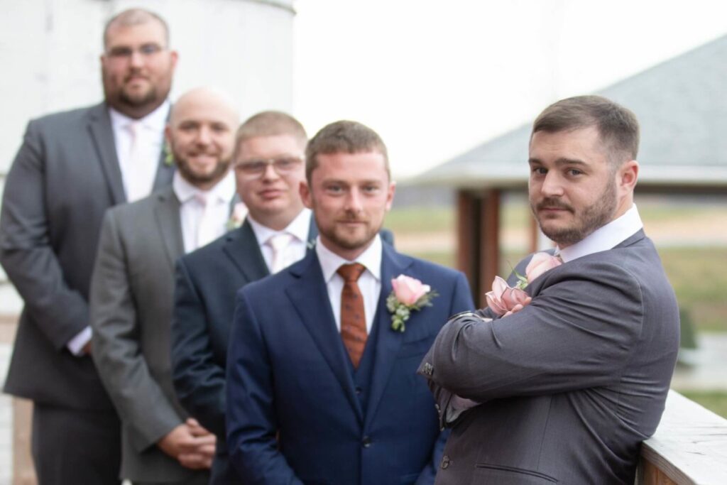 Christine-Jon-Wedding-Blog-61 - TOMMY GATZ ENTERTAINMENT Five men in suits stand in a line outdoors. The man in front, wearing a gray suit, crosses his arms. Each man has a boutonniere, with the central figure in a navy suit.