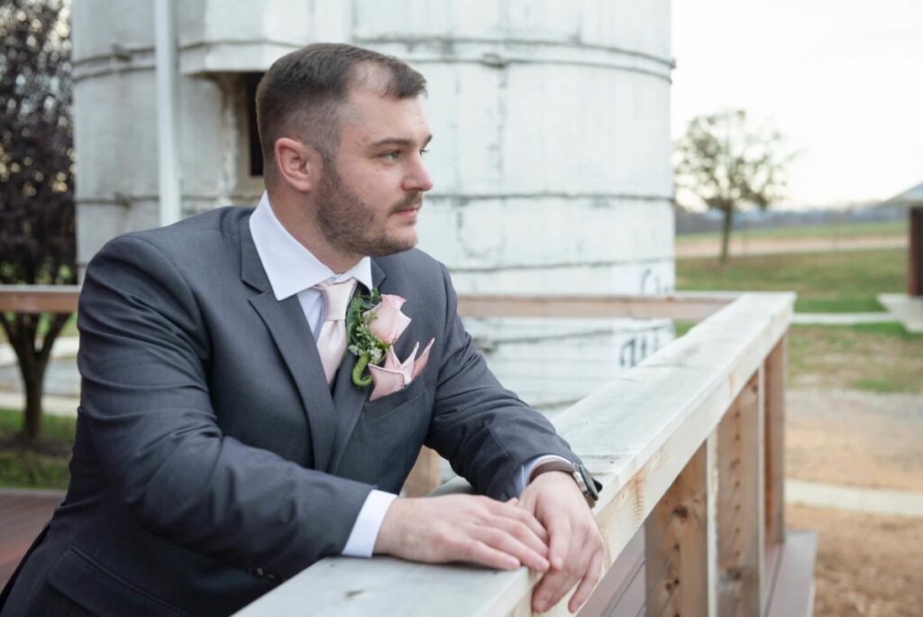 Christine-Jon-Wedding-Blog-62 - TOMMY GATZ ENTERTAINMENT A man in a suit with a boutonniere stands by a wooden railing, looking into the distance. Background includes a silo and some trees.