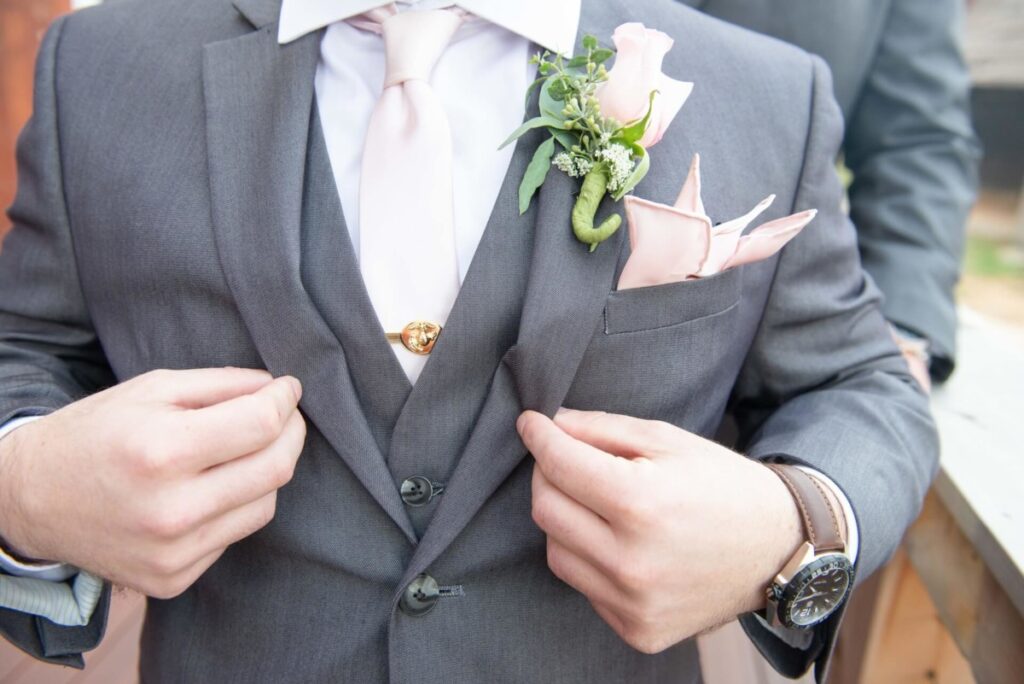 Christine-Jon-Wedding-Blog-63 - TOMMY GATZ ENTERTAINMENT Man in a gray suit adjusts his jacket, wearing a pink tie and boutonniere, with a watch on his wrist and a pink pocket square.