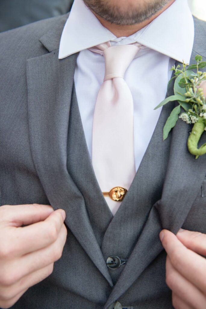 Christine-Jon-Wedding-Blog-64 - TOMMY GATZ ENTERTAINMENT A man in a gray suit adjusts his jacket. He wears a light pink tie and a gold tie clip. A boutonniere with greenery and a pink flower is on his left lapel.
