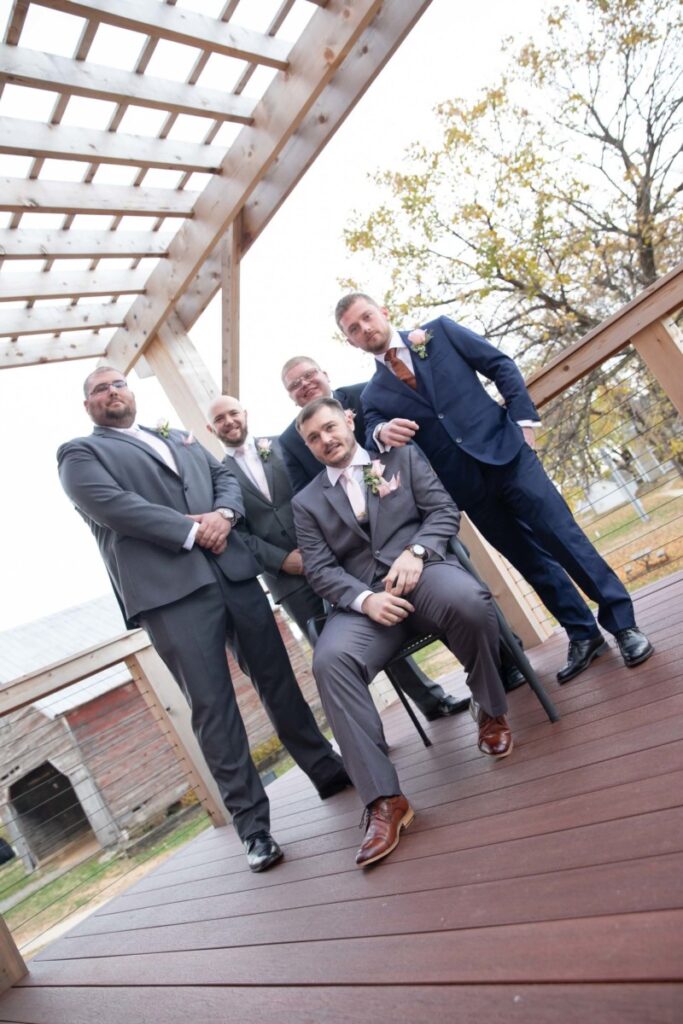 Christine-Jon-Wedding-Blog-65 - TOMMY GATZ ENTERTAINMENT Five men in suits, one sitting on a chair, pose under a wooden pergola on a deck. Trees and a barn are in the background.