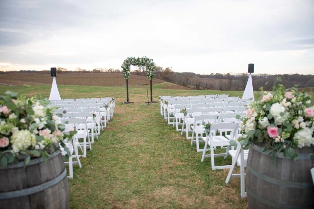 Christine-Jon-Wedding-Blog-66 - TOMMY GATZ ENTERTAINMENT An outdoor wedding ceremony setup with white chairs arranged in rows, a floral arch at the front, and flower arrangements on wine barrels. The setting is a grassy field with a scenic backdrop.