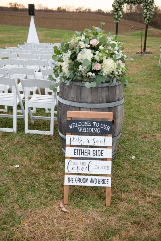 Christine-Jon-Wedding-Blog-67 - TOMMY GATZ ENTERTAINMENT Outdoor wedding setup with white chairs and floral arrangements. A wooden sign reads, "Welcome to our wedding. Pick a seat either side. You are loved by both the groom and bride.
