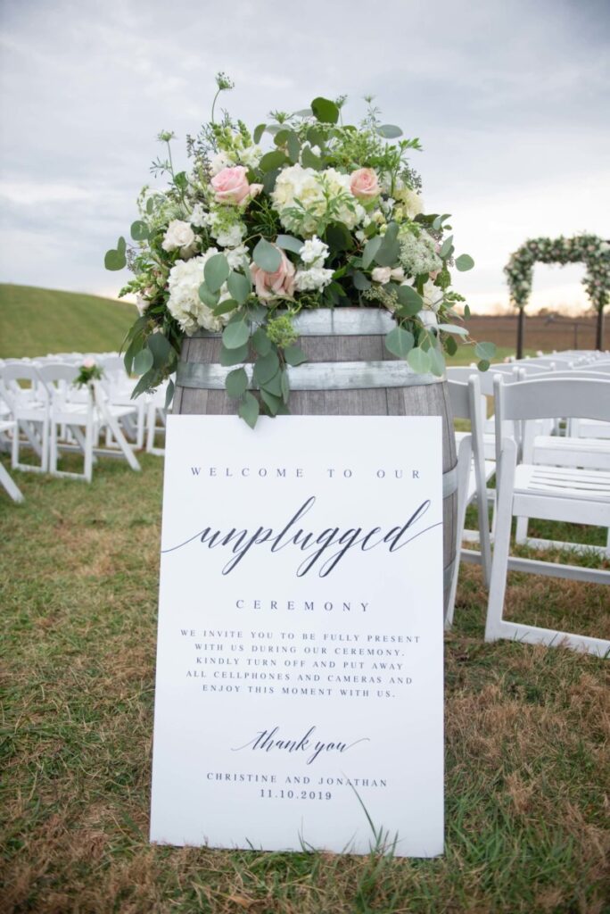 Christine-Jon-Wedding-Blog-68 - TOMMY GATZ ENTERTAINMENT A wooden barrel with a floral arrangement and a sign reading "Welcome to our unplugged ceremony" is placed among rows of white chairs outdoors on a grassy area.