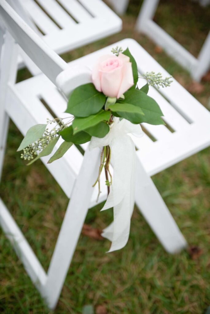 Christine-Jon-Wedding-Blog-69 - TOMMY GATZ ENTERTAINMENT A white folding chair on grass, decorated with a pink rose, green leaves, and a white ribbon.