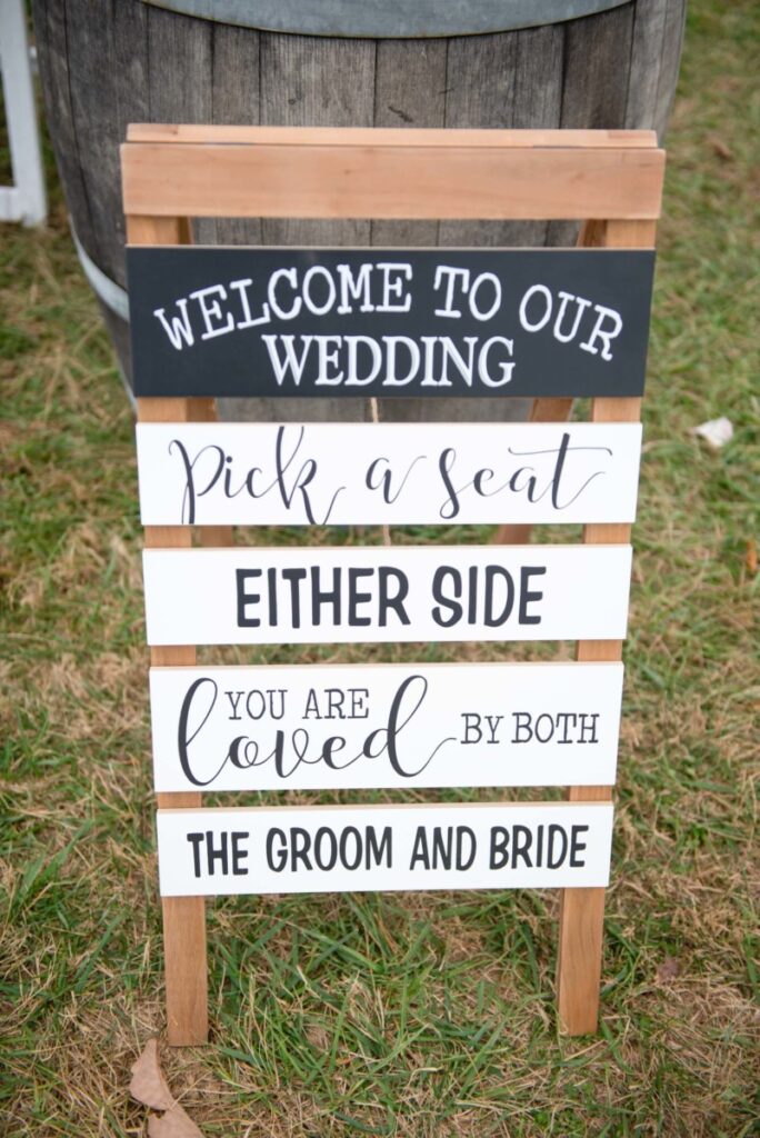 Christine-Jon-Wedding-Blog-70 - TOMMY GATZ ENTERTAINMENT Wooden wedding sign on grass reads: "Welcome to our wedding. Pick a seat, either side. You are loved by both the groom and bride.