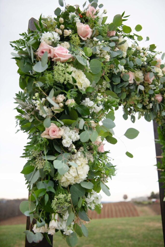 Christine-Jon-Wedding-Blog-72 - TOMMY GATZ ENTERTAINMENT Floral arrangement with pink roses, white hydrangeas, and greenery on a wooden arch outdoors.