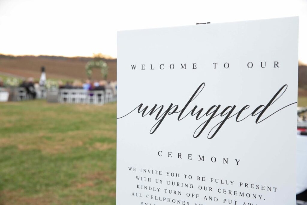 Christine-Jon-Wedding-Blog-73 - TOMMY GATZ ENTERTAINMENT Sign at an outdoor wedding reads, "Welcome to our unplugged ceremony," requesting guests turn off devices.