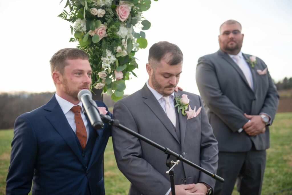 Christine-Jon-Wedding-Blog-74 - TOMMY GATZ ENTERTAINMENT Three men in suits stand outdoors at a microphone, with floral decorations behind them. Two men have serious expressions, while one looks down.