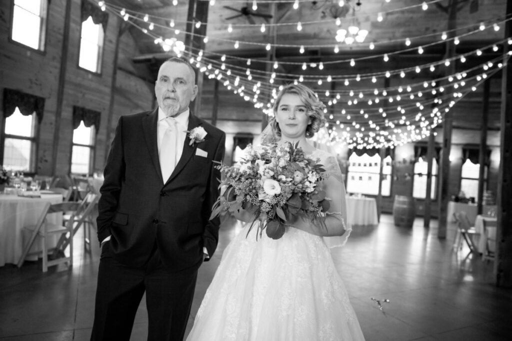 Christine-Jon-Wedding-Blog-75 - TOMMY GATZ ENTERTAINMENT A bride and an older gentleman walk down the aisle in a decorated venue with string lights. She holds a bouquet, and both are dressed formally.