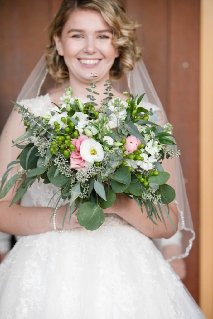 Christine-Jon-Wedding-Blog-76 - TOMMY GATZ ENTERTAINMENT Bride smiling and holding a bouquet with green leaves, white flowers, and pink accents, wearing a white lace wedding dress and veil, standing in front of a wooden background.