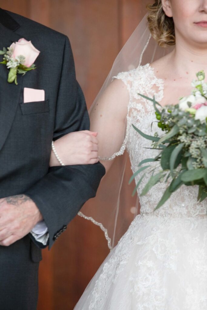 Christine-Jon-Wedding-Blog-77 - TOMMY GATZ ENTERTAINMENT A bride in a lace dress holds a bouquet and links arms with a groom in a dark suit, both wearing floral corsages.