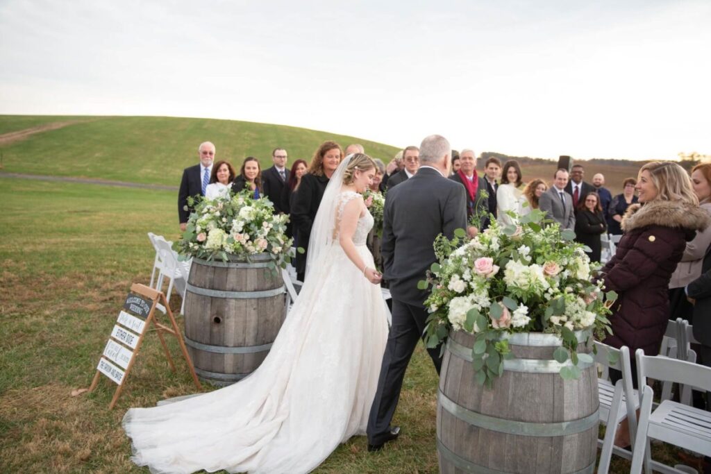 Christine-Jon-Wedding-Blog-78 - TOMMY GATZ ENTERTAINMENT Bride in a white gown walks down an outdoor aisle adorned with flowers and barrels, accompanied by an older man. Guests stand on either side, watching the procession.
