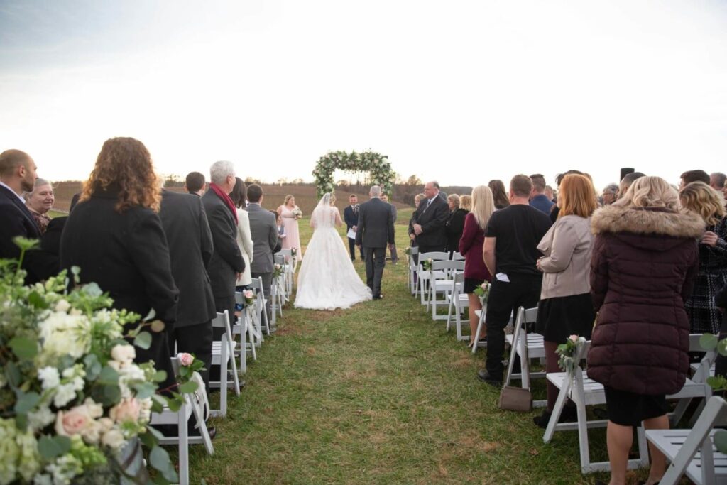 Christine-Jon-Wedding-Blog-79 - TOMMY GATZ ENTERTAINMENT Bride walks down an outdoor aisle with guests seated on either side, towards an arch adorned with greenery and flowers.