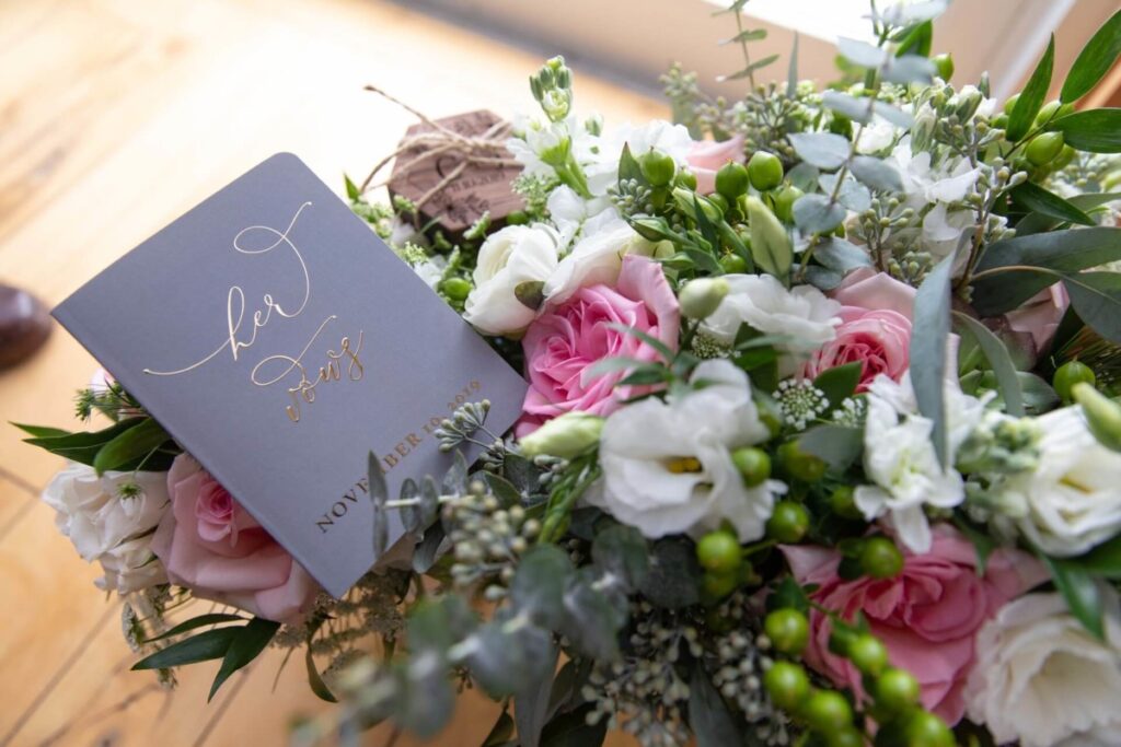 Christine-Jon-Wedding-Blog-8 - TOMMY GATZ ENTERTAINMENT A bouquet of pink and white flowers with greenery is placed next to a gray card titled "her vows.
