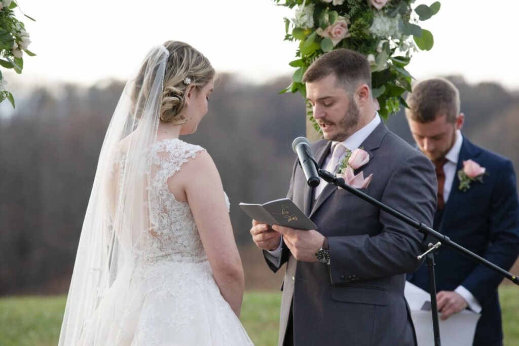 Christine-Jon-Wedding-Blog-80 - TOMMY GATZ ENTERTAINMENT A groom reads vows from a notebook at an outdoor wedding ceremony. The bride, in a lace gown, listens. A groomsman stands in the background, holding papers. Floral decorations are visible.