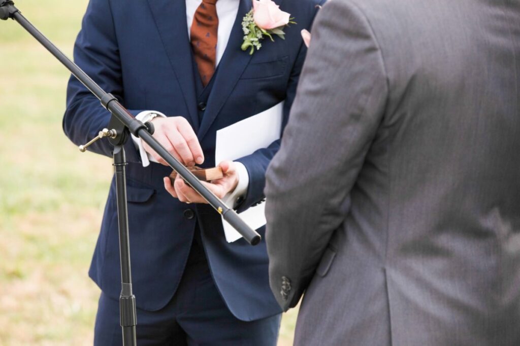 Christine-Jon-Wedding-Blog-81 - TOMMY GATZ ENTERTAINMENT Two people in suits are standing near a microphone. One holds a paper and a phone. A boutonniere is pinned to his jacket.