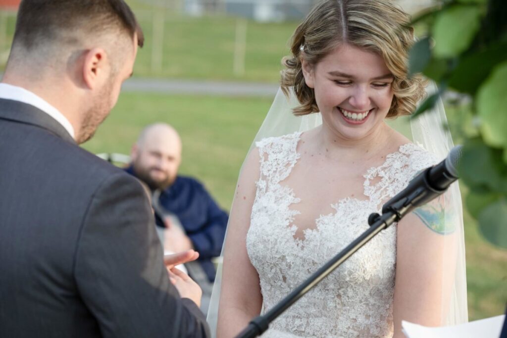 Christine-Jon-Wedding-Blog-82 - TOMMY GATZ ENTERTAINMENT Bride smiling as groom places ring during outdoor wedding ceremony. Person in background watching.
