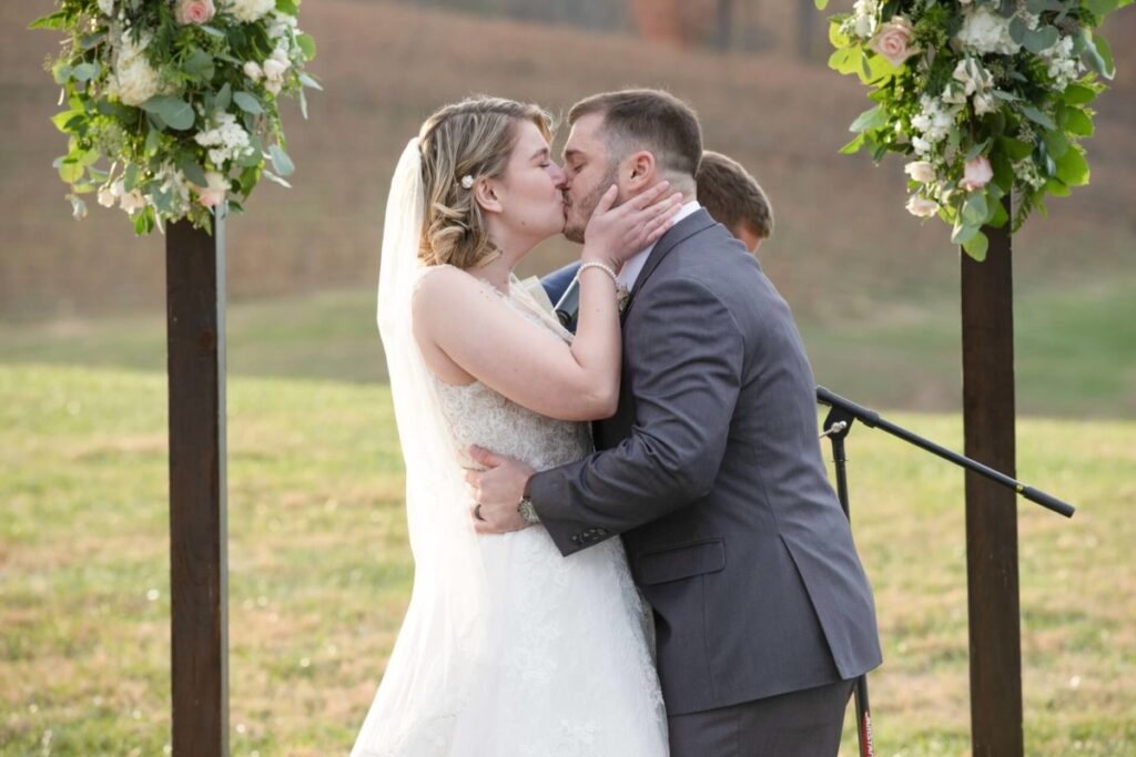 Christine-Jon-Wedding-Blog-83 - TOMMY GATZ ENTERTAINMENT A bride and groom kiss under a floral arch during an outdoor wedding ceremony.