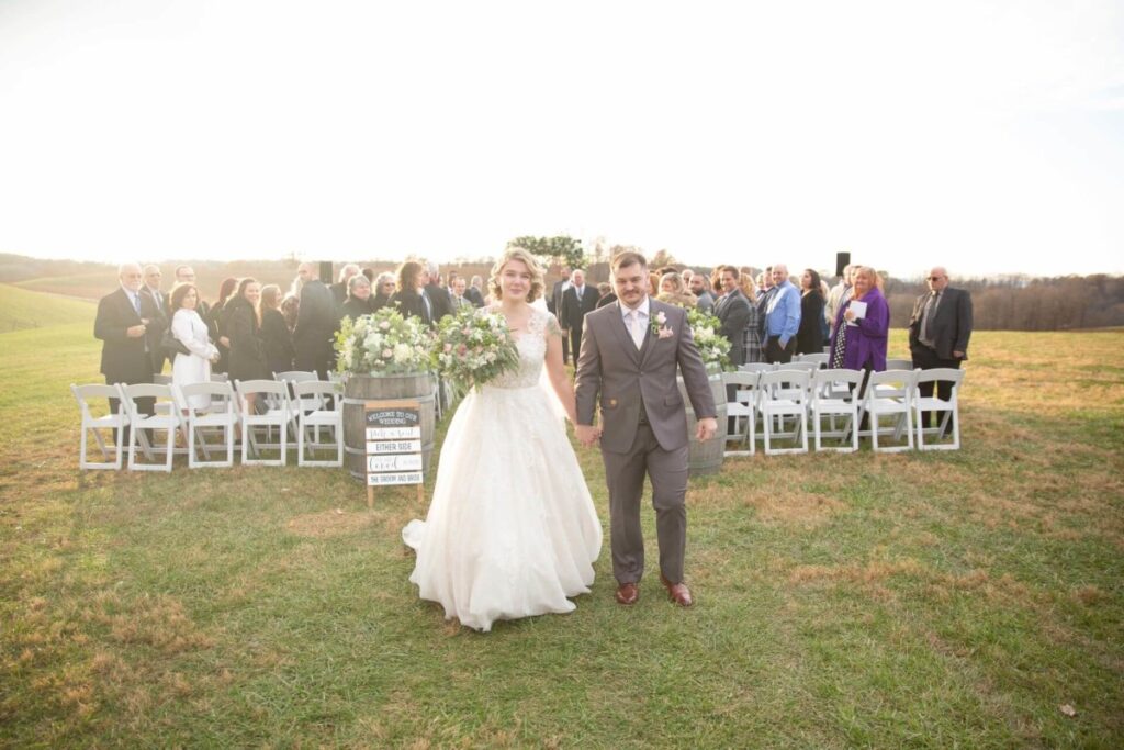 Christine-Jon-Wedding-Blog-84 - TOMMY GATZ ENTERTAINMENT A bride in a white gown and a groom in a gray suit walk hand in hand outdoors. A group of people in formal attire is seated behind them, and a sign stands near the aisle.