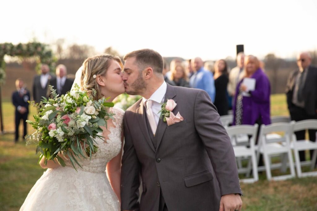 Christine-Jon-Wedding-Blog-85 - TOMMY GATZ ENTERTAINMENT A couple kisses outdoors during a wedding ceremony. The bride holds a bouquet, and guests are visible in the background.