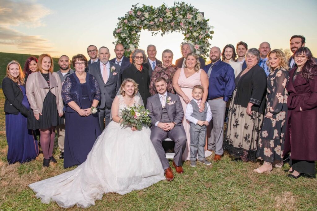 Christine-Jon-Wedding-Blog-86 - TOMMY GATZ ENTERTAINMENT Bride and groom surrounded by a large group of family and friends outdoors, standing on grass in front of a floral arch.