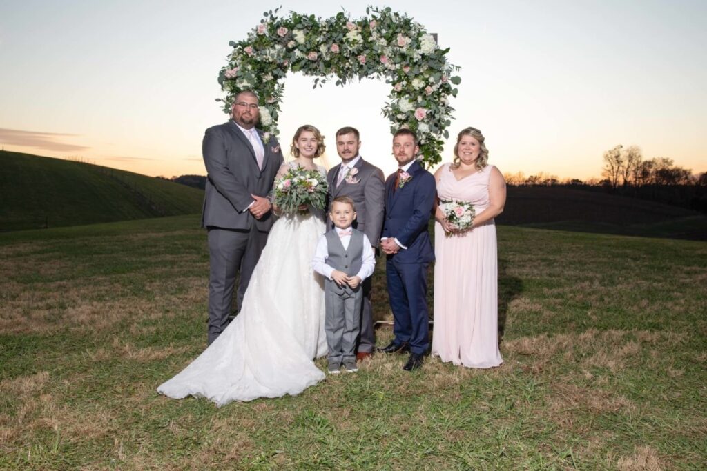 Christine-Jon-Wedding-Blog-87 - TOMMY GATZ ENTERTAINMENT A wedding party stands in front of a flower arch outdoors at sunset. The group includes the bride, groom, a young boy, and three adults, all dressed in formal attire.
