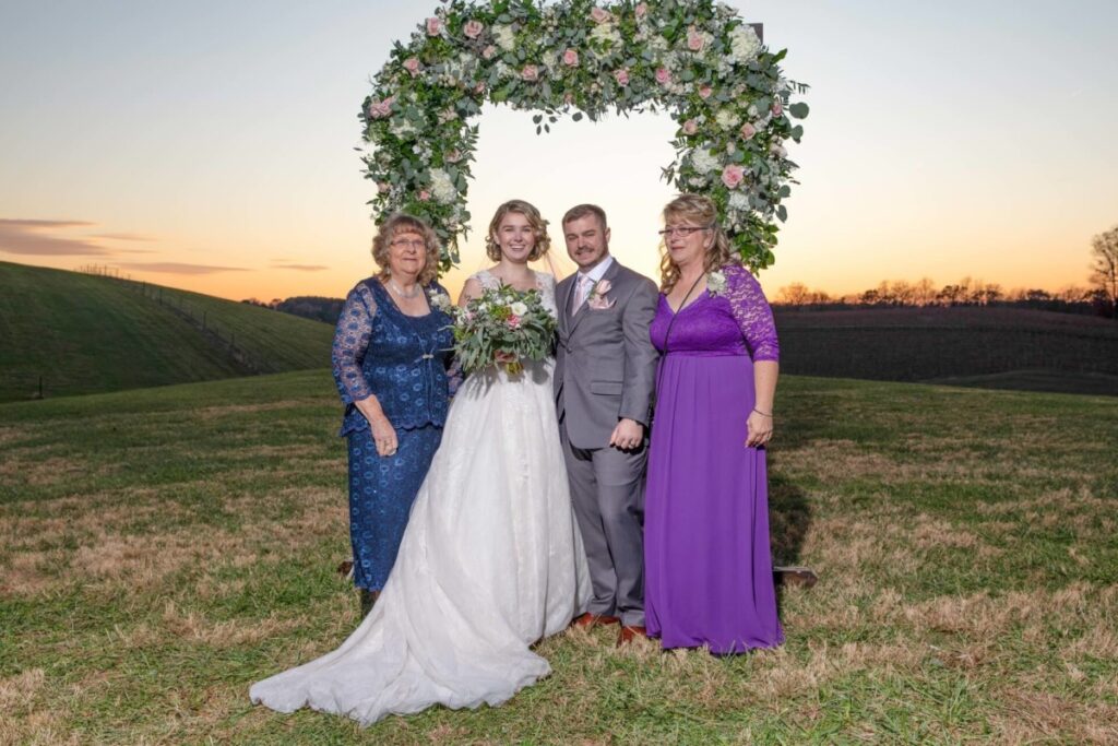 Christine-Jon-Wedding-Blog-88 - TOMMY GATZ ENTERTAINMENT A bride and groom pose with two women under a floral arch at sunset on a grassy hill.