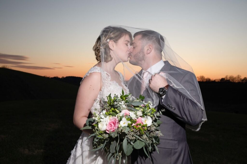 Christine-Jon-Wedding-Blog-89 - TOMMY GATZ ENTERTAINMENT A bride and groom kiss under a veil at sunset, holding a bouquet with pink and white flowers.