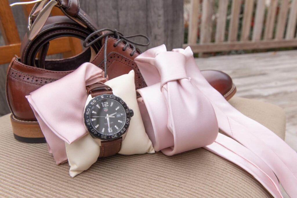Christine-Jon-Wedding-Blog-9 - TOMMY GATZ ENTERTAINMENT Brown leather shoes, a belt, three pink satin ties, and a watch with a brown strap. The items are arranged on a beige surface with a wooden fence in the background.