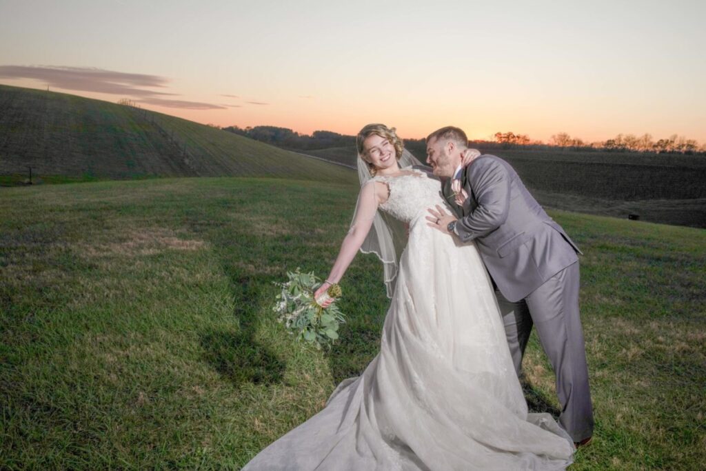 Christine-Jon-Wedding-Blog-90 - TOMMY GATZ ENTERTAINMENT A couple in wedding attire poses playfully outdoors on a grassy field at sunset. The bride holds a bouquet and leans backward, while the groom supports her.