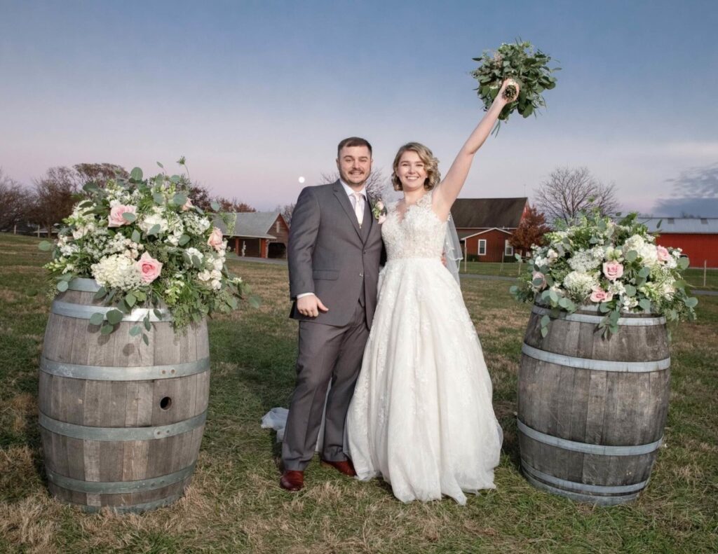 Christine-Jon-Wedding-Blog-91 - TOMMY GATZ ENTERTAINMENT A couple poses outdoors on their wedding day, standing between two floral arrangements on barrels. The bride holds a bouquet up in celebration.