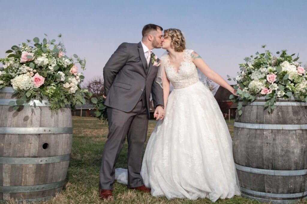 Christine-Jon-Wedding-Blog-92 - TOMMY GATZ ENTERTAINMENT A bride and groom kiss outdoors between two floral arrangements on barrels, with a grassy field and a clear sky in the background.